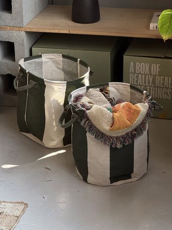 The OYOY Living Design Kara Storage Basket Medium, Set of 2, features wide green and white stripes. One basket holds a colorful woven blanket and orange items, while the other is empty. Both sit on a gray floor with boxes and shelves in the background.