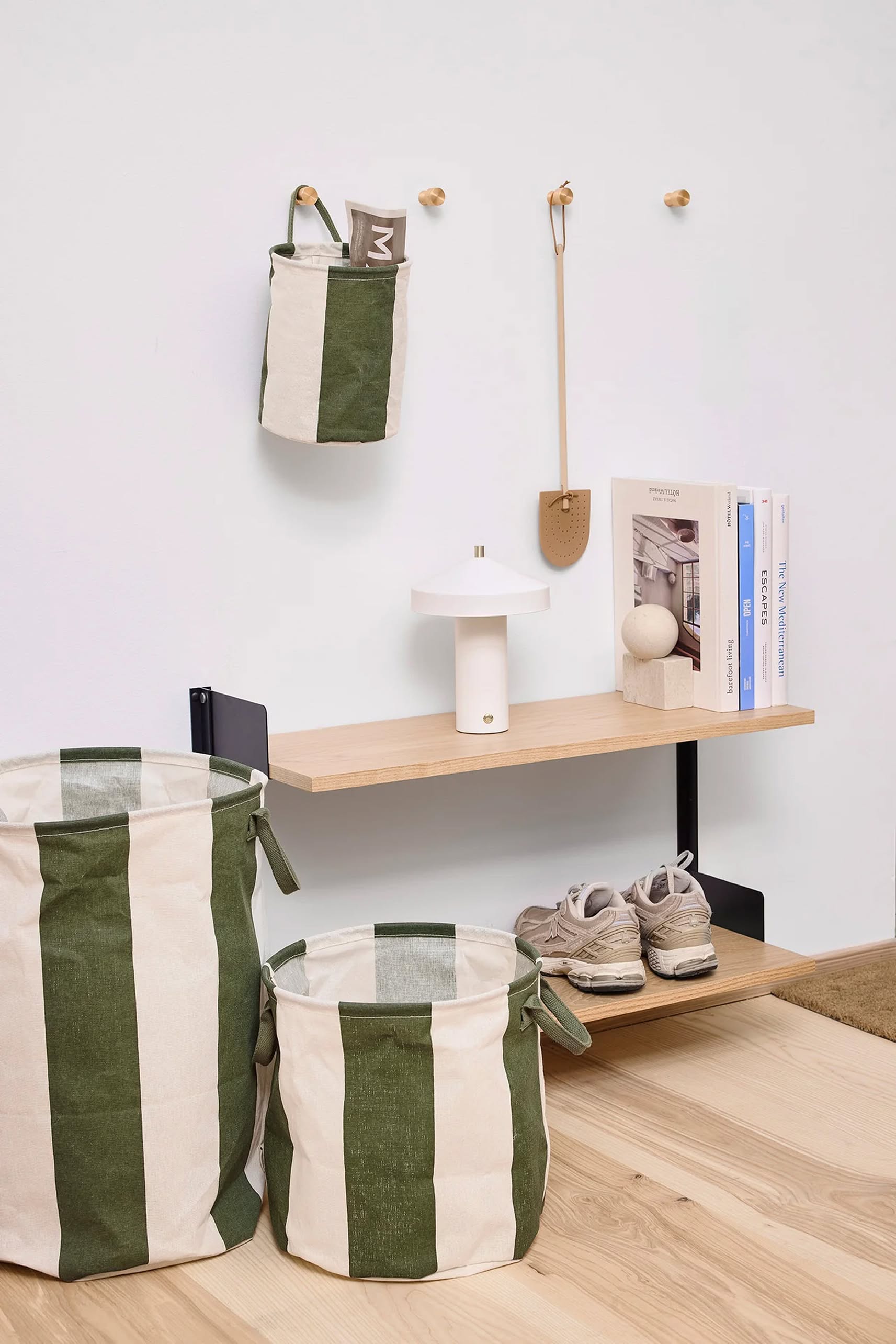 A modern entryway features OYOY Living Design Kara Storage Basket Medium, Set of 2, in green and white stripes with PE-coated interiors for easy organizing, plus a shelf with books, a white lamp, beige sneakers, and a hanging bag and brush above light wood flooring.