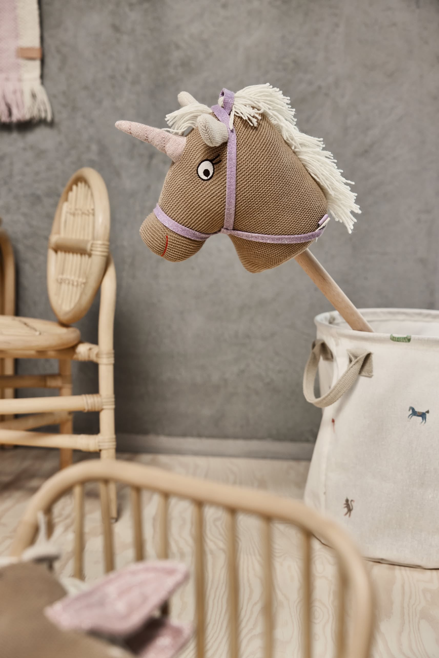 A fabric unicorn hobby horse with a pink horn and white mane peeks from an OYOY Living Design Moira Laundry & Storage Basket (Medium) in a cozy, neutral kids’ room with wicker furniture and wooden floors.