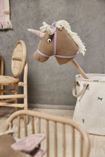 A fabric unicorn hobby horse with a pink horn and white mane peeks from an OYOY Living Design Moira Laundry & Storage Basket (Medium) in a cozy, neutral kids’ room with wicker furniture and wooden floors.