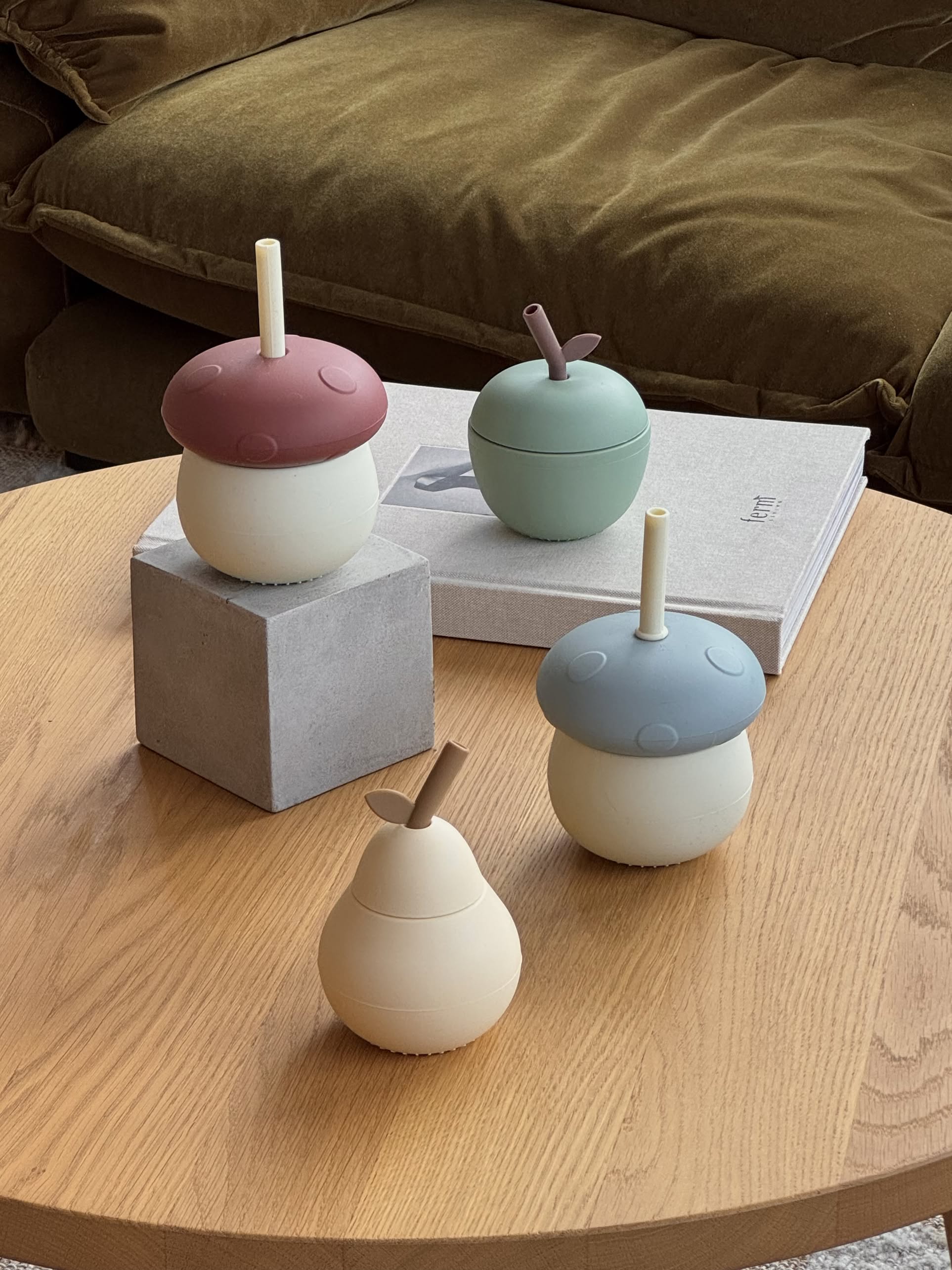 Four playful OYOY Living Design Mushroom Cups, each with silicone lids and straws, are arranged on a wooden table—two placed on books and a gray cube—while a brown sofa in the background adds cozy charm.