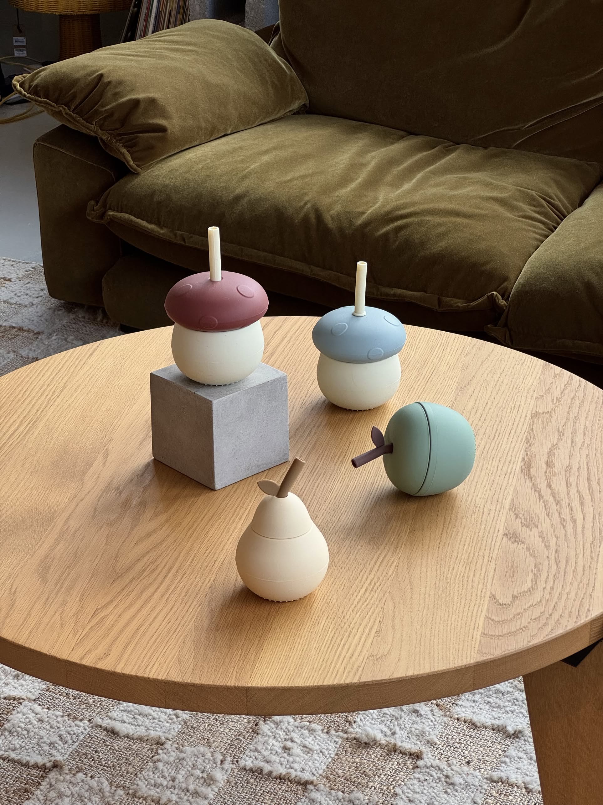 Four playful OYOY Living Design Mushroom Cups with straws, in soft pastel colors and whimsical fruit-inspired shapes, are displayed on a wooden coffee table in front of a green couch.