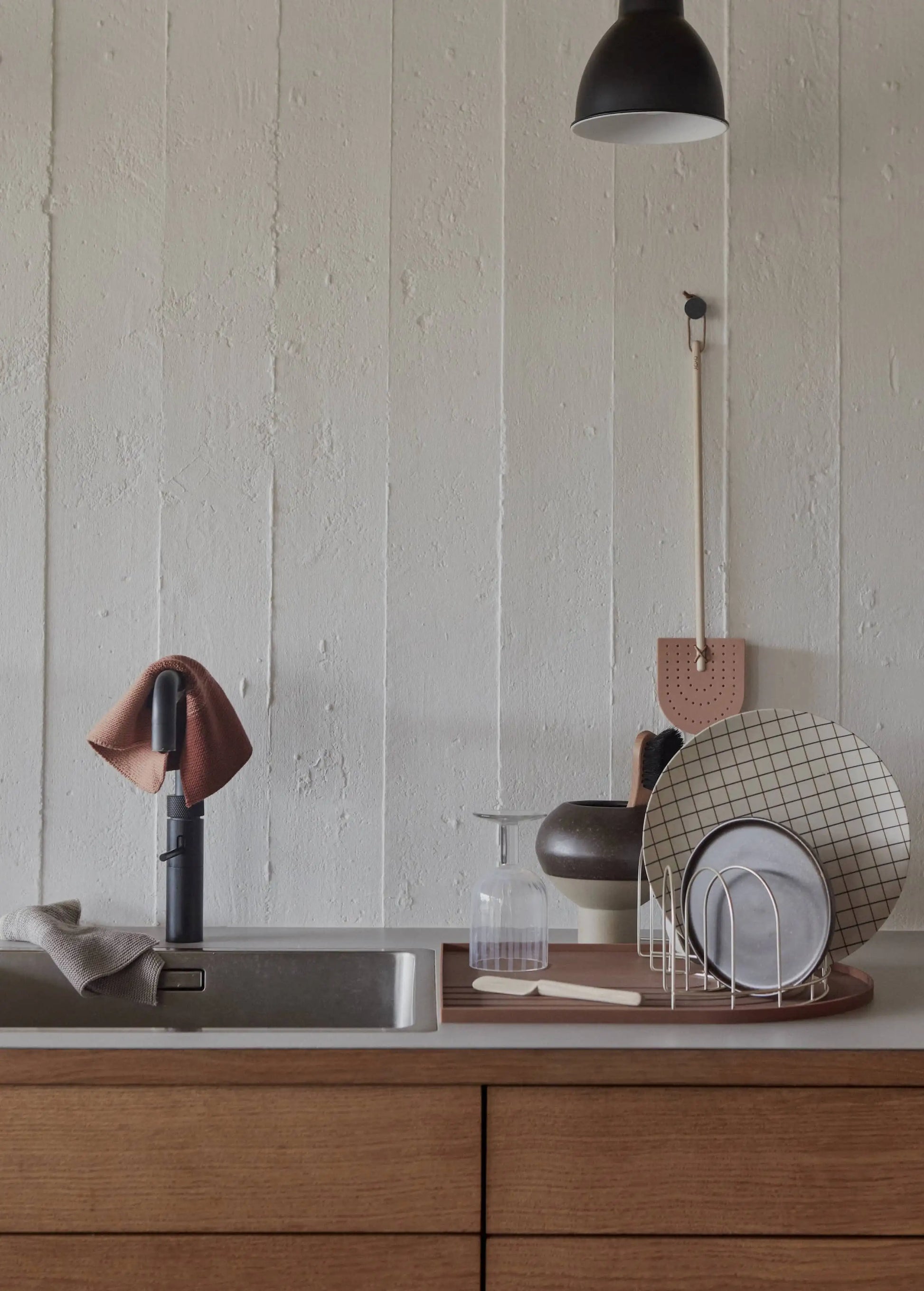 A minimalist kitchen sink setup features OYOY Living Design Niji Dish Cloths draped over the faucet, a beige hand towel, drying rack with plates, a soap dispenser, and pendant lighting against a textured off-white wall.