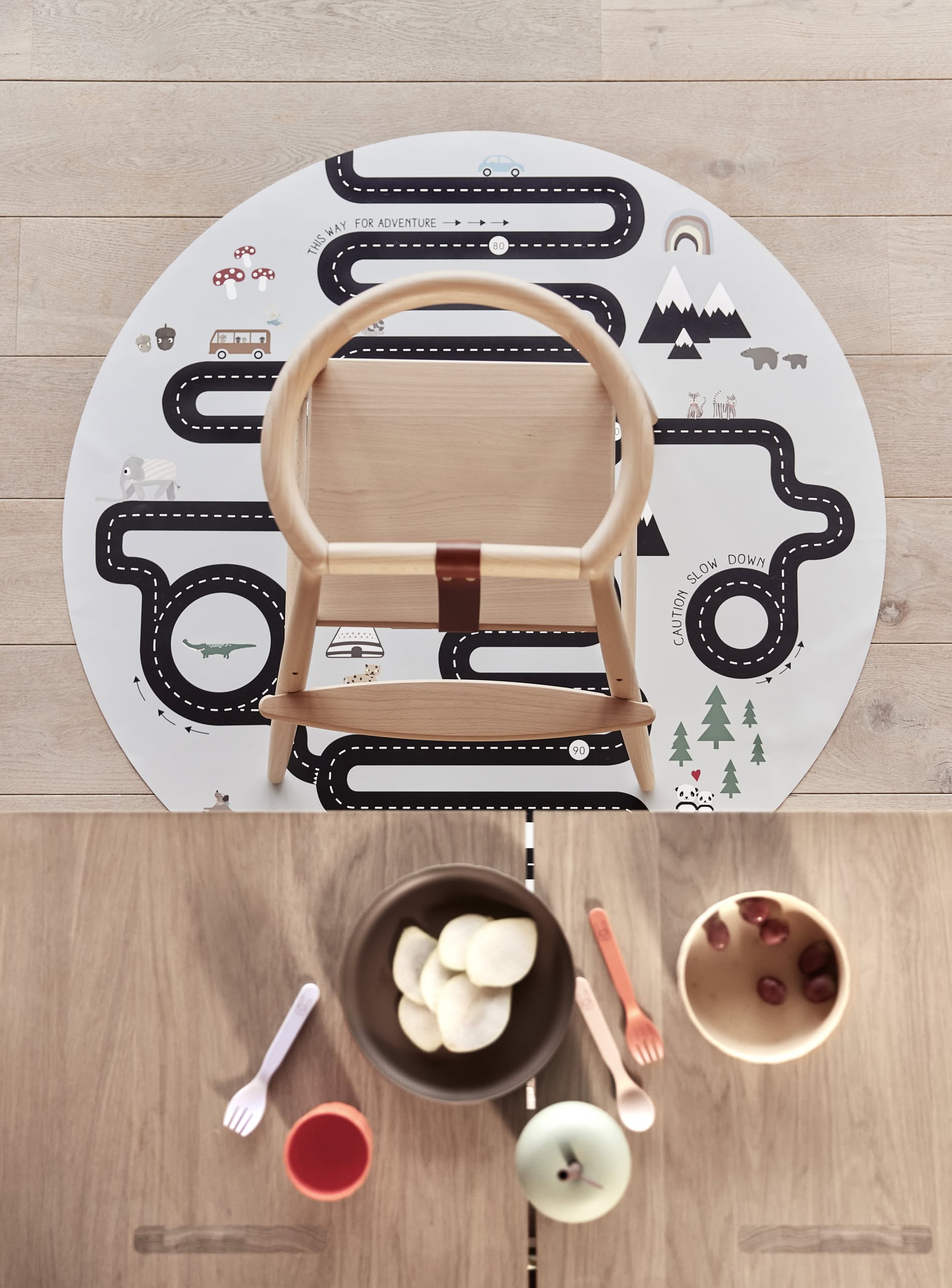 A top-down view of a wooden high chair on a round road-patterned play mat atop light wood floors. On the table, bowls of fruit and the OYOY Living Design Pullo Cutlery Set by OYOY Living Design add vibrant color.