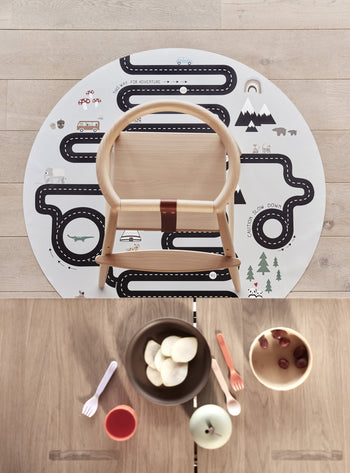 A top-down view of a wooden high chair on a round road-patterned play mat atop light wood floors. On the table, bowls of fruit and the OYOY Living Design Pullo Cutlery Set by OYOY Living Design add vibrant color.