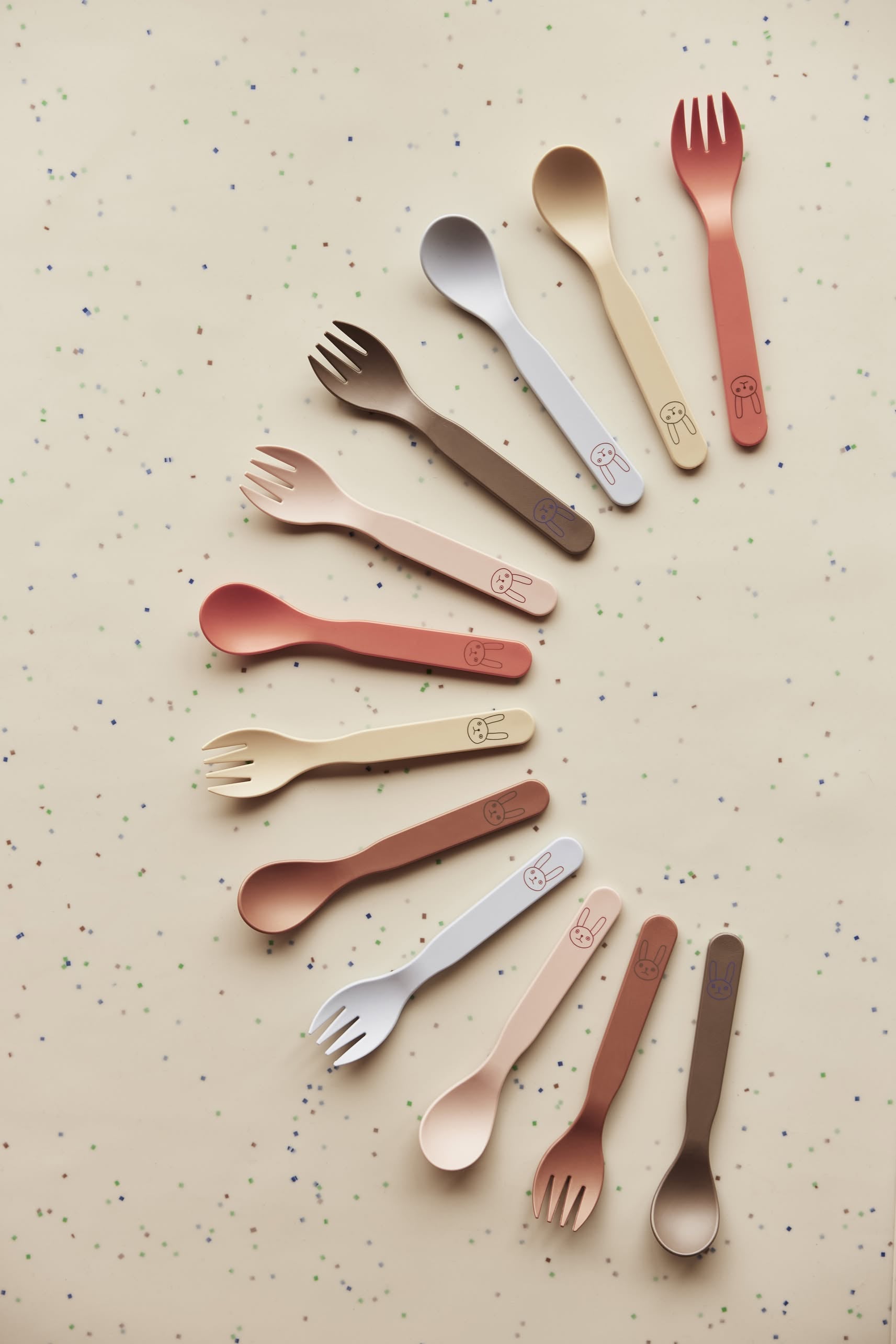 The OYOY Living Design Pullo Cutlery Set features colorful, eco-friendly PLA sporks and spoons arranged in a spiral on a beige, dotted surface.