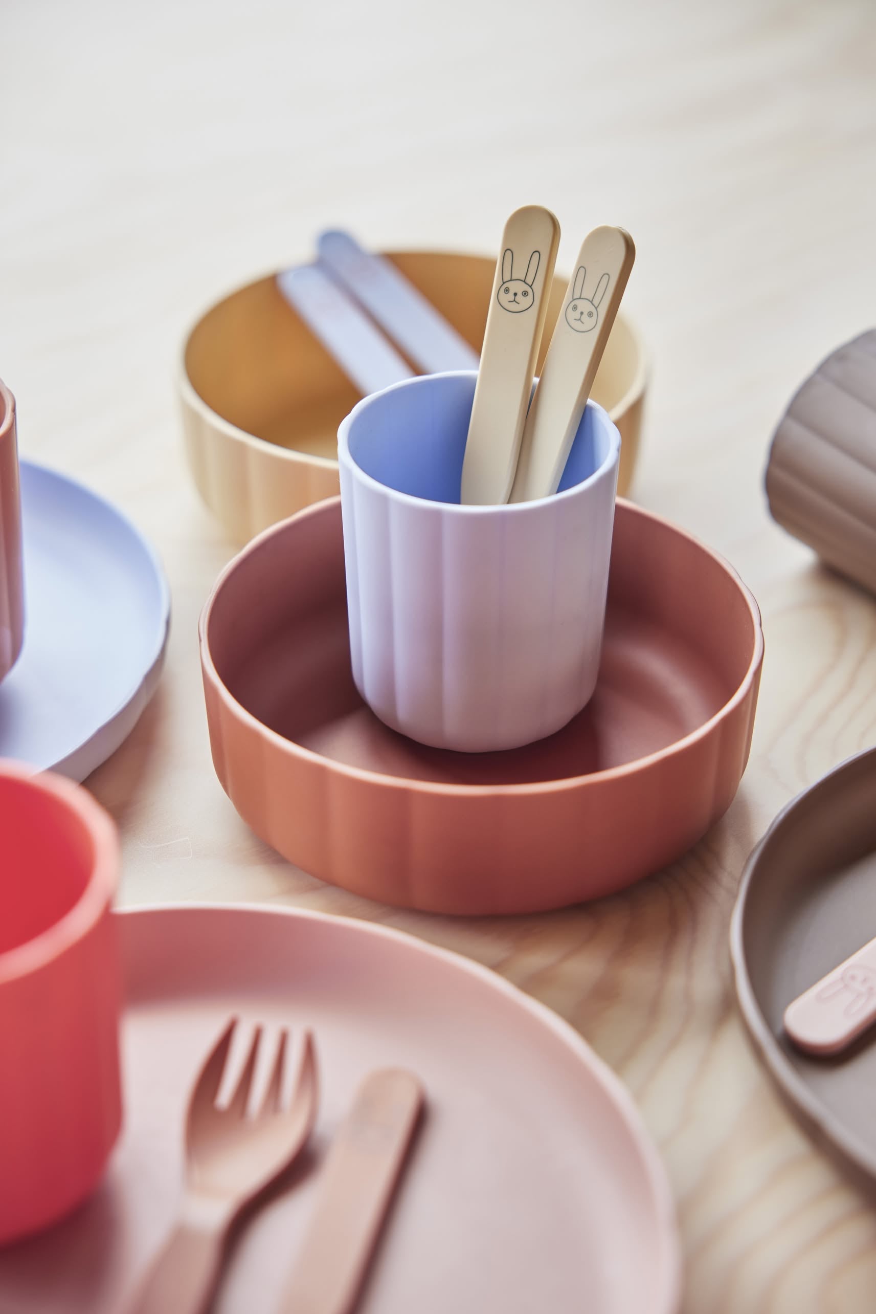 Pastel-colored plates, bowls, and cups from OYOY Living Design’s Pullo collection, made with eco-friendly PLA, are arranged on light wood. Two beige OYOY Living Design Pullo spoons with bunny faces complete this adorable toddler cutlery set.