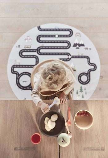 A young child uses the OYOY Living Design Pullo Cutlery Set at a wooden table with bowls of apples and grapes, a red cup, and a playmat featuring a road design placed on the floor behind them.
