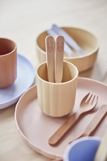 The OYOY Living Design Pullo Cutlery Set features pastel-colored, eco-friendly PLA children’s dishware—including a pink plate, tan cup, bunny-face spoons, and matching fork and spoon—beautifully displayed on a light wooden surface.