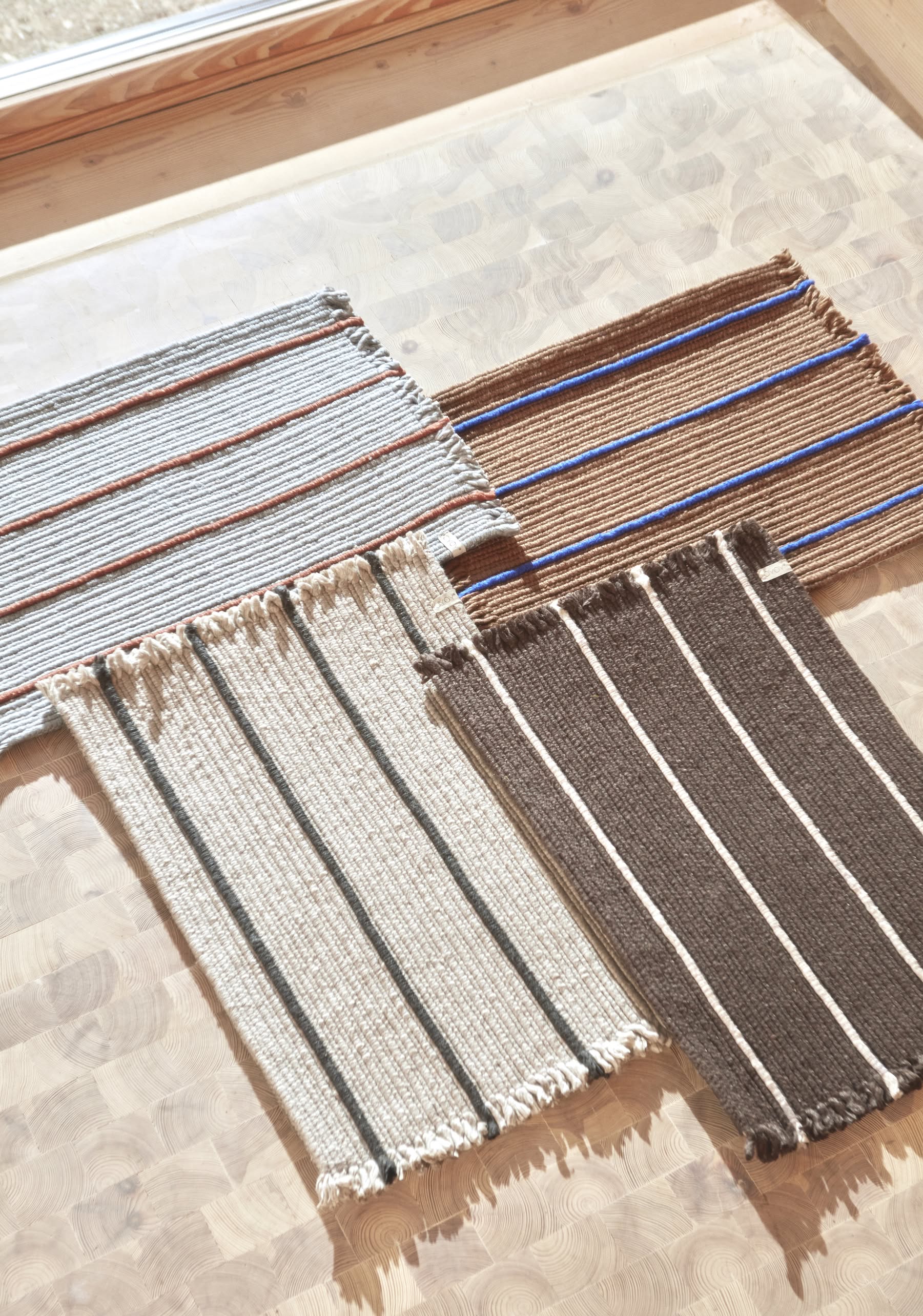Four OYOY Living Design Putki Recycled Doormats with fringe, featuring simple horizontal stripes in beige, brown, cream, and blue, are displayed on a light wooden surface in natural sunlight.