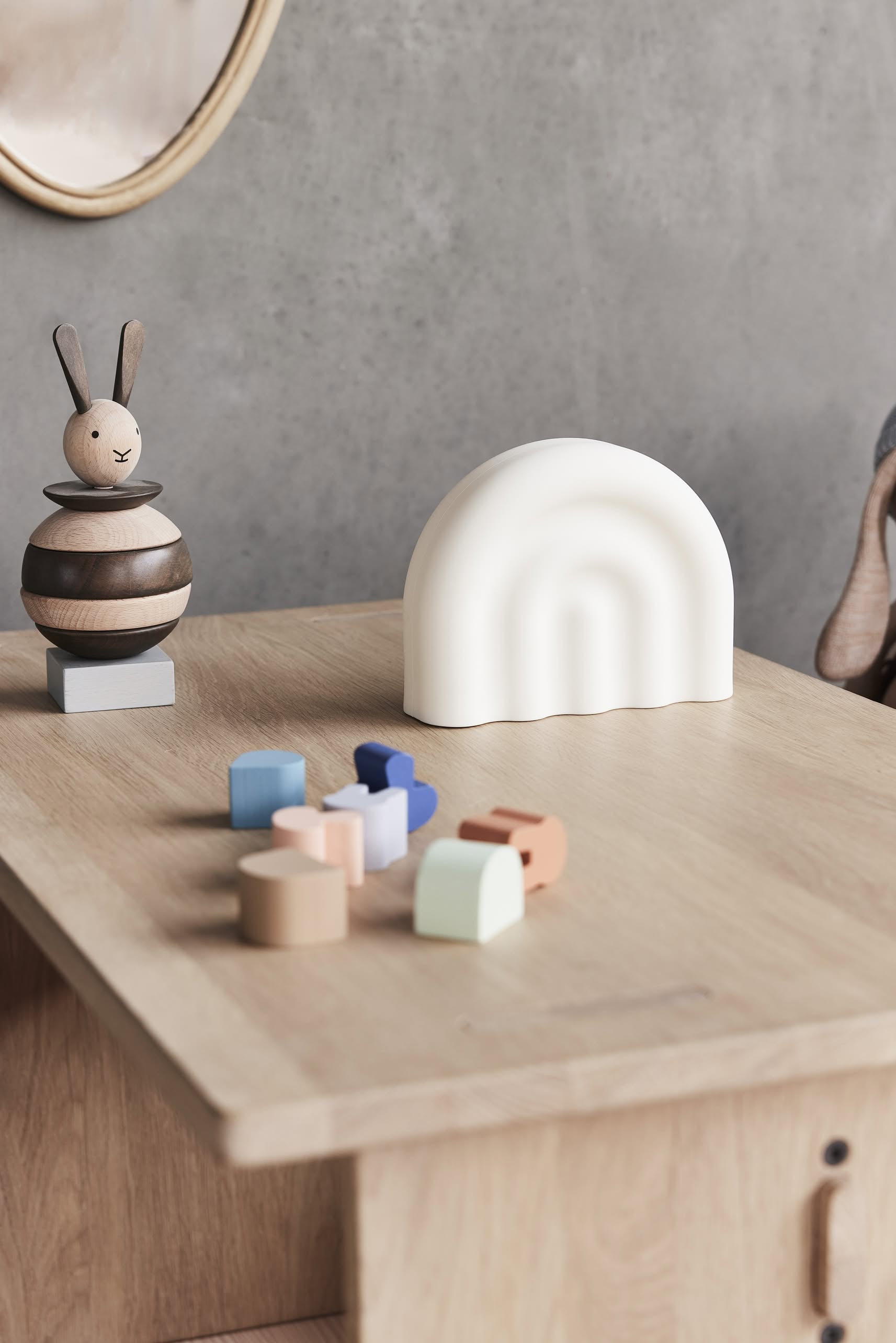 A wooden desk displays a wooden bunny toy, the OYOY Living Design Rainbow Night Light in a soft silicone white arc shape, and colorful geometric blocks. The background includes a gray wall and part of another wooden object.