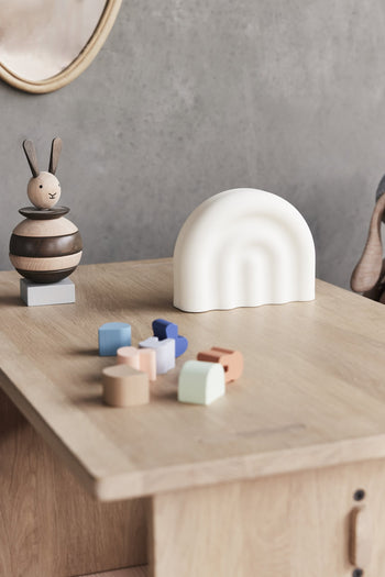 A wooden desk displays a wooden bunny toy, the OYOY Living Design Rainbow Night Light in a soft silicone white arc shape, and colorful geometric blocks. The background includes a gray wall and part of another wooden object.