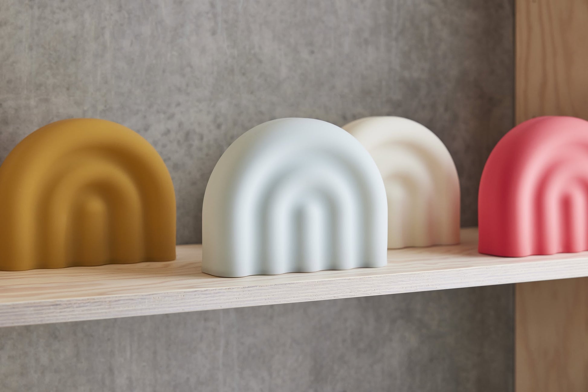 Four rounded, arch-shaped OYOY Living Design Rainbow Night Lights in mustard yellow, pale blue, off-white, and coral pink glow softly on a light wooden shelf against a gray concrete wall.
