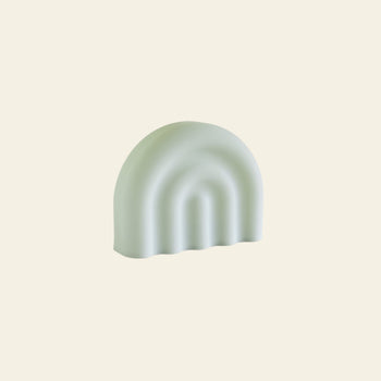 A pale green, dome-shaped soft silicone object with three arched grooves and a wavy base edge sits on a light beige background, showcasing the modern, minimalist style of the OYOY Living Design Rainbow Night Light by OYOY Living Design.