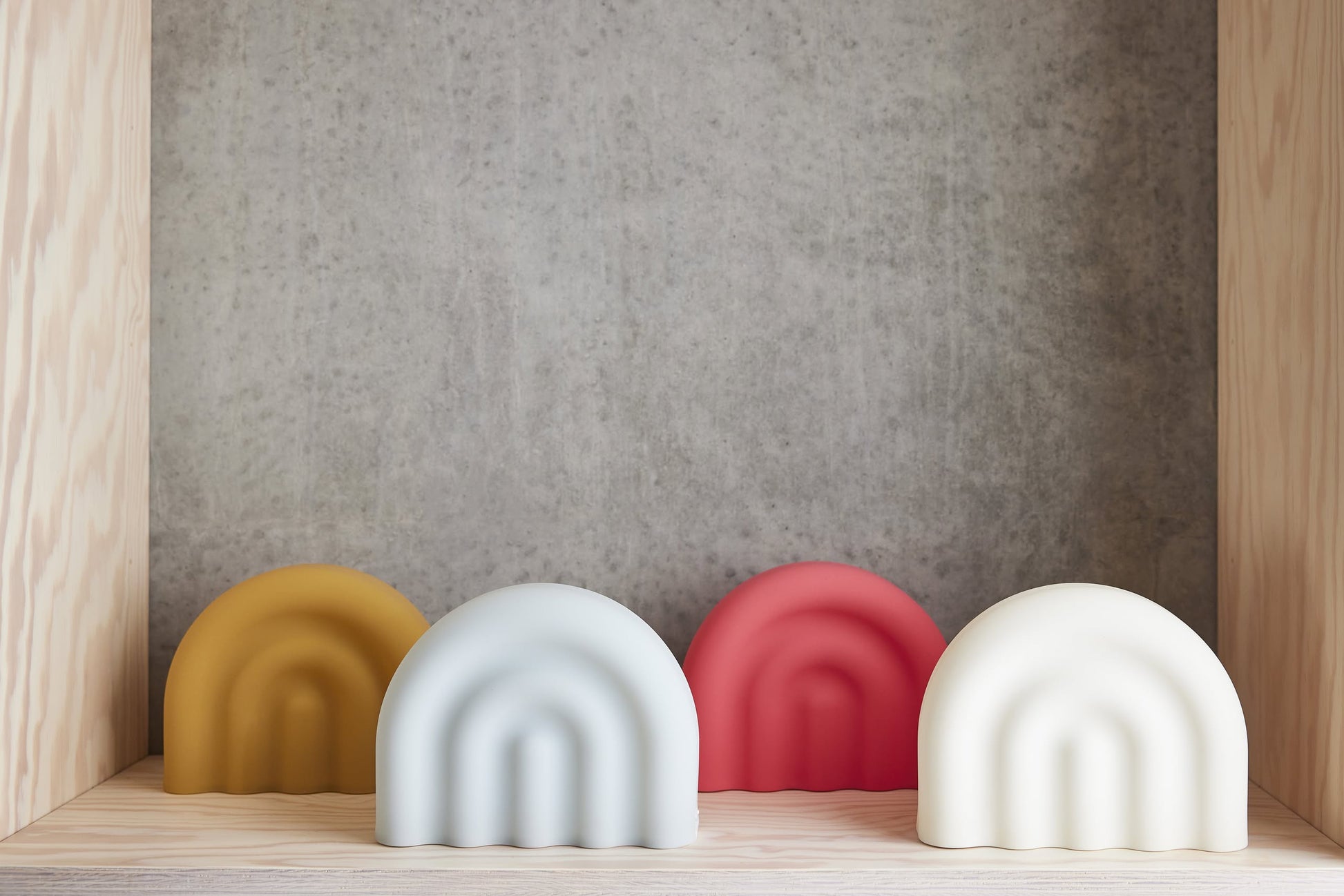 Four arch-shaped OYOY Living Design Rainbow Night Lights in yellow, light blue, red, and white—crafted from soft silicone with a minimalist design—sit on a wooden shelf, glowing softly against a concrete backdrop.