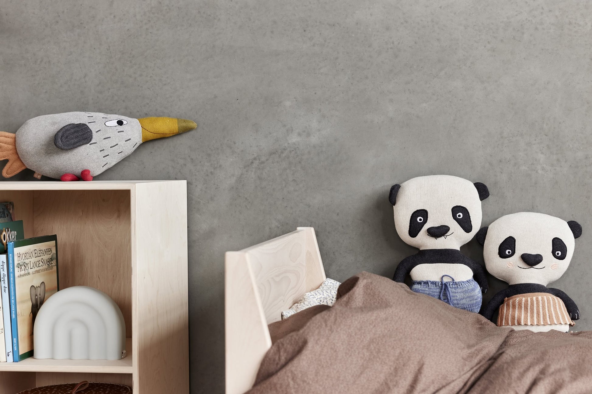 Stuffed pandas sit on a bed with brown bedding by a gray wall; a plush bird with a yellow beak rests on a light wood bookshelf featuring books and the OYOY Living Design Rainbow Night Light, which casts a soothing glow.