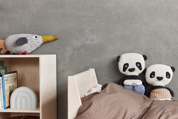Stuffed pandas sit on a bed with brown bedding by a gray wall; a plush bird with a yellow beak rests on a light wood bookshelf featuring books and the OYOY Living Design Rainbow Night Light, which casts a soothing glow.