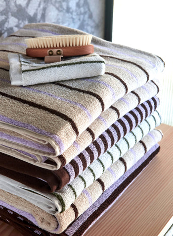 A neatly folded stack of OYOY Living Design Raita Wash Cloths (Pack of 2) sits on a wooden surface, accented by two small brushes. Sunlight streams in, creating a calm, tidy atmosphere.