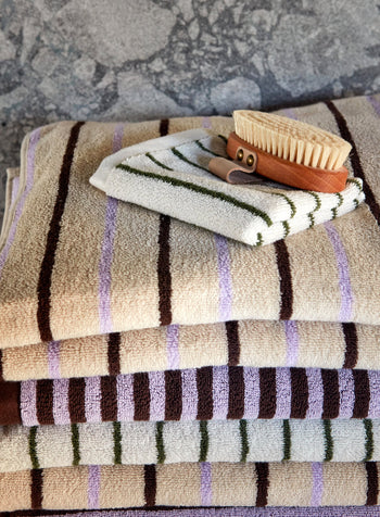 A stack of striped OYOY Living Design Raita Wash Cloths in beige, brown, and lavender sits on a surface, topped with an organic cotton Terry wash cloth and a wooden bath brush with natural bristles.