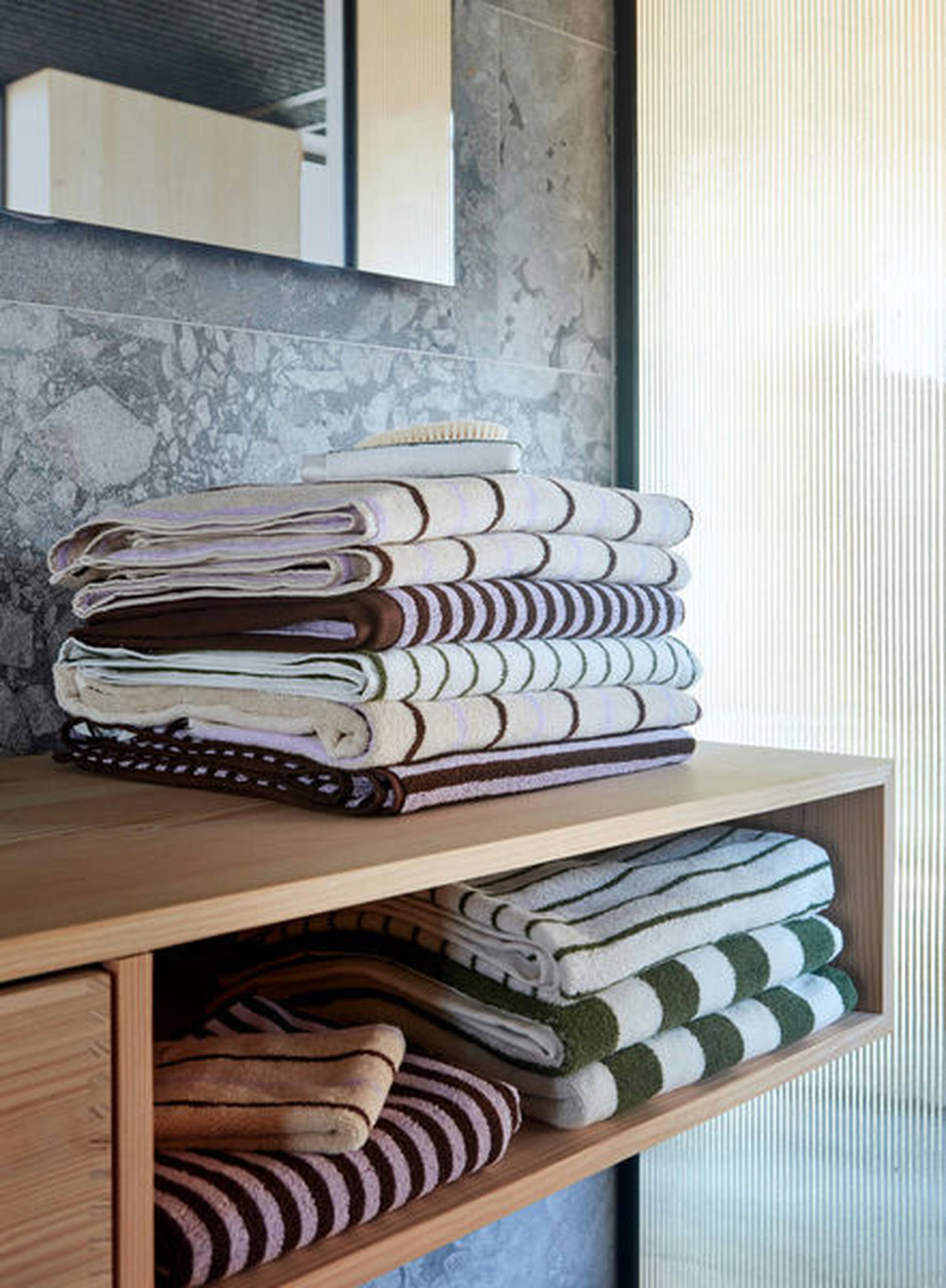 A pack of OYOY Living Design Raita Wash Cloths, made of organic cotton in green, brown, and white, rests on a wooden shelf in a modern bathroom with stone walls and a translucent panel.
