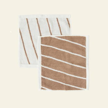 The OYOY Living Design Raita Wash Cloth, Pack of 2, features organic cotton terry cloths with diagonal stripes—one mainly white with slim brown stripes, one mainly brown with slim white stripes—each bordered in white on a light beige background.