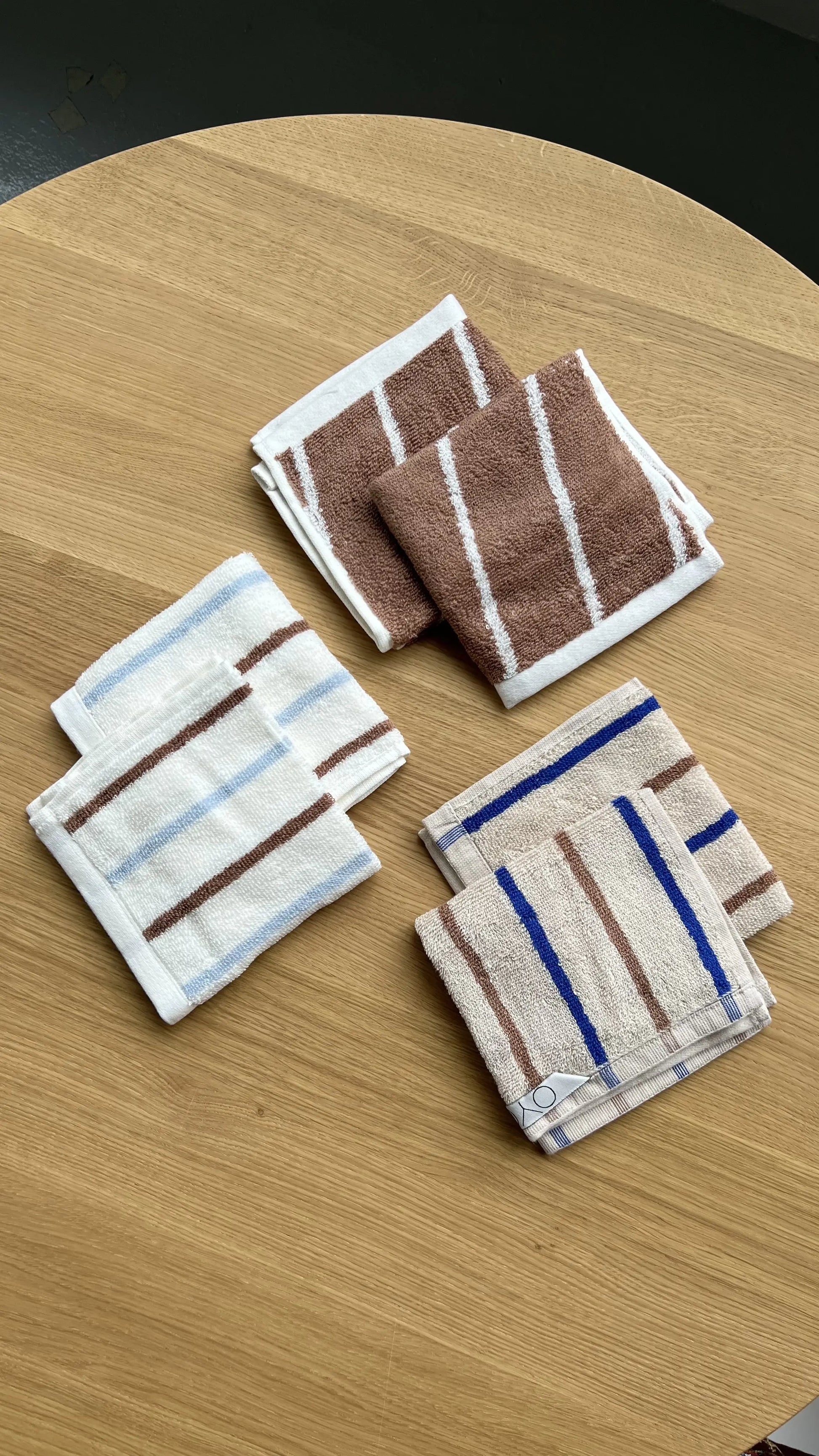 Three stacks of OYOY Living Design Raita Wash Cloths, Pack of 2, made from organic cotton terry with striped patterns in brown/white, white/brown, and tan/blue accents, are displayed on a light wooden surface.