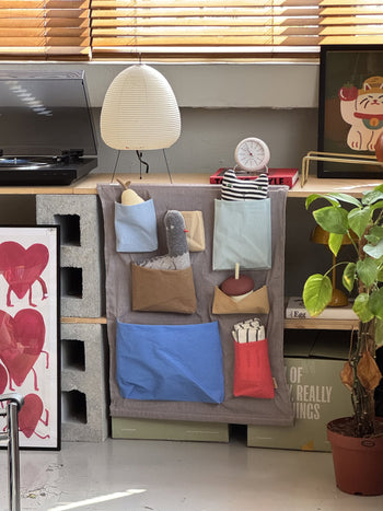 A cozy corner features the OYOY Living Design Saga Wall Storage from OYOY Living Design, neatly holding stuffed items, a record player, white clock, cat figurine, and potted plant as sunlight shines through wooden blinds.