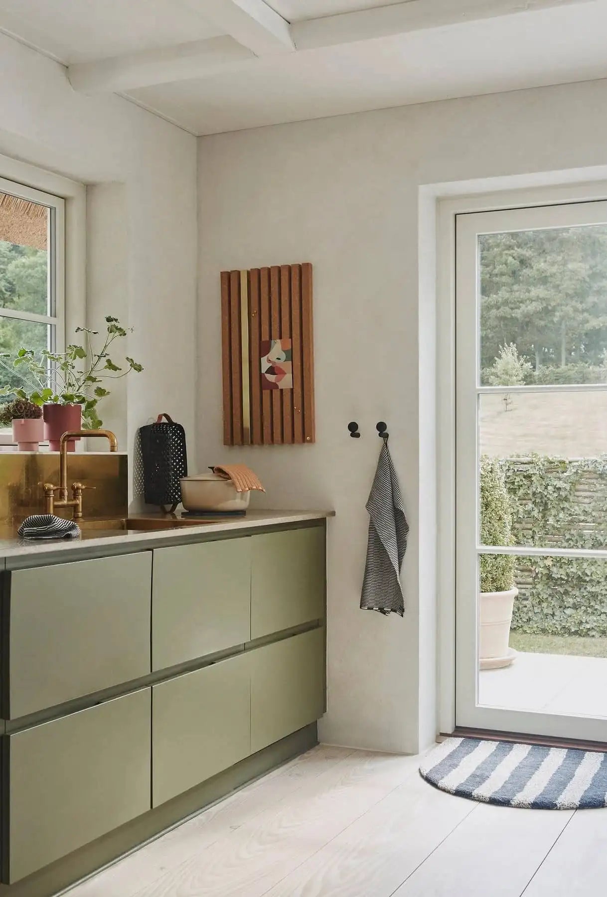 A modern kitchen with olive green cabinets, gold sink, potted plants, and a dish rack displays OYOY Living Design Stringa Mini Towels hanging near a glass door by wall hooks. A striped rug is placed by the entrance.