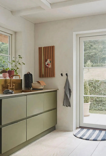 A modern kitchen with olive green cabinets, gold sink, potted plants, and a dish rack displays OYOY Living Design Stringa Mini Towels hanging near a glass door by wall hooks. A striped rug is placed by the entrance.