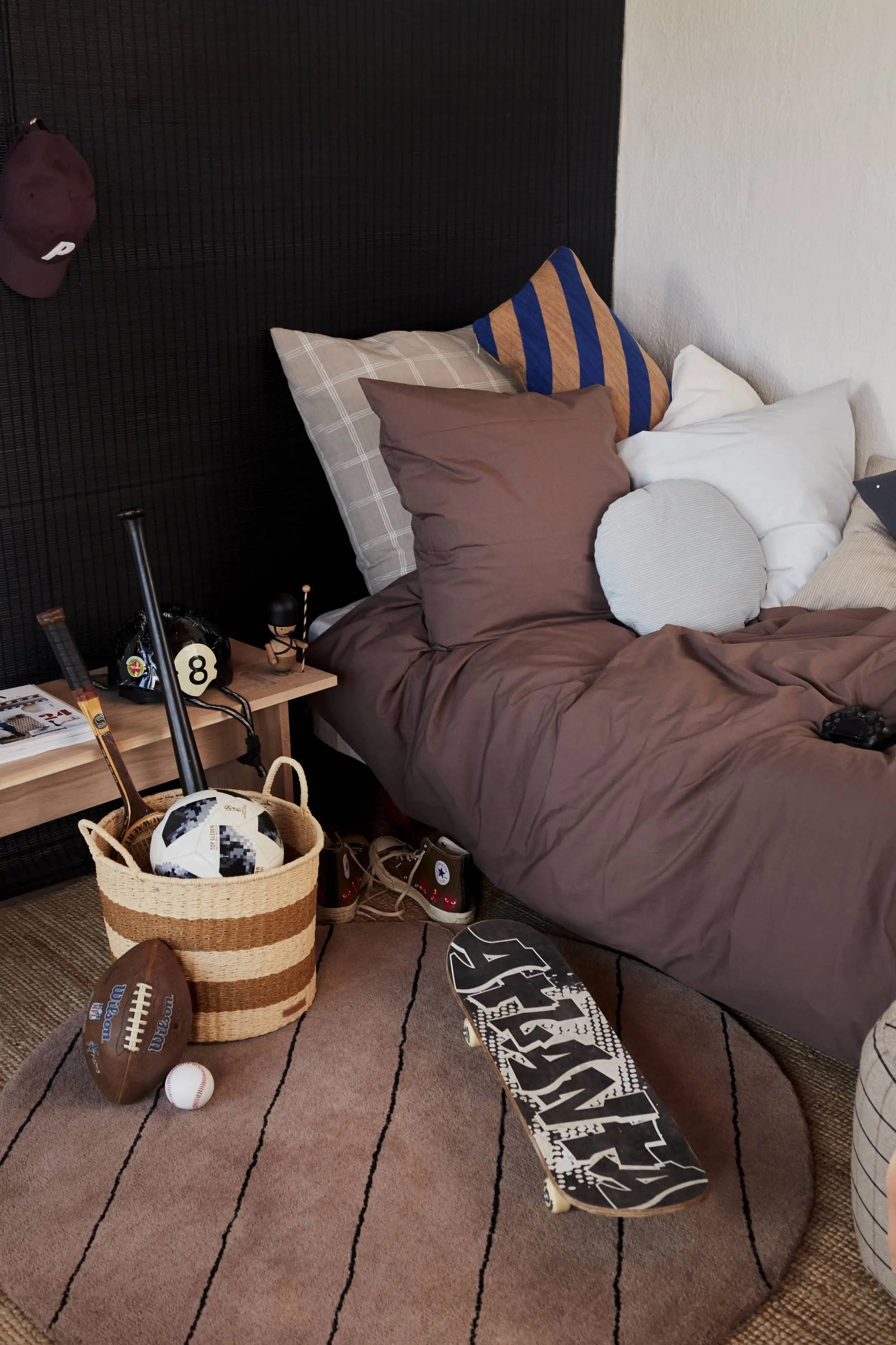 A cozy bedroom features a brown bed, assorted pillows, and the OYOY Living Design Striped Circle Rug in wool-cotton. Sports gear sits on the bedside table; a basket with bats, basketball, and football is by a skateboard—great for sporty home decor.