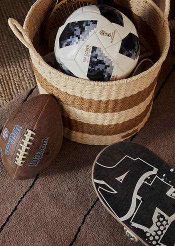 A striped woven basket holds a black-and-white soccer ball. Nearby, an American football and a graphic skateboard rest on the OYOY Living Design Striped Circle Rug—perfect for blending decor styles.