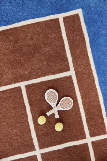 Two small white toy tennis rackets and two yellow balls rest atop the OYOY Living Design Tennis Rug, crafted from a wool-cotton blend and designed by OYOY Living Design to resemble a sporty brown and blue court with crisp white boundary lines.