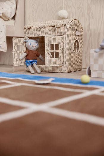 A gray stuffed animal in a brown shirt and blue striped shorts sits in the dollhouse doorway. In the foreground, the OYOY Living Design Tennis Rug by OYOY Living Design, crafted from a wool-cotton blend, adds sporty flair with a yellow ball nearby.