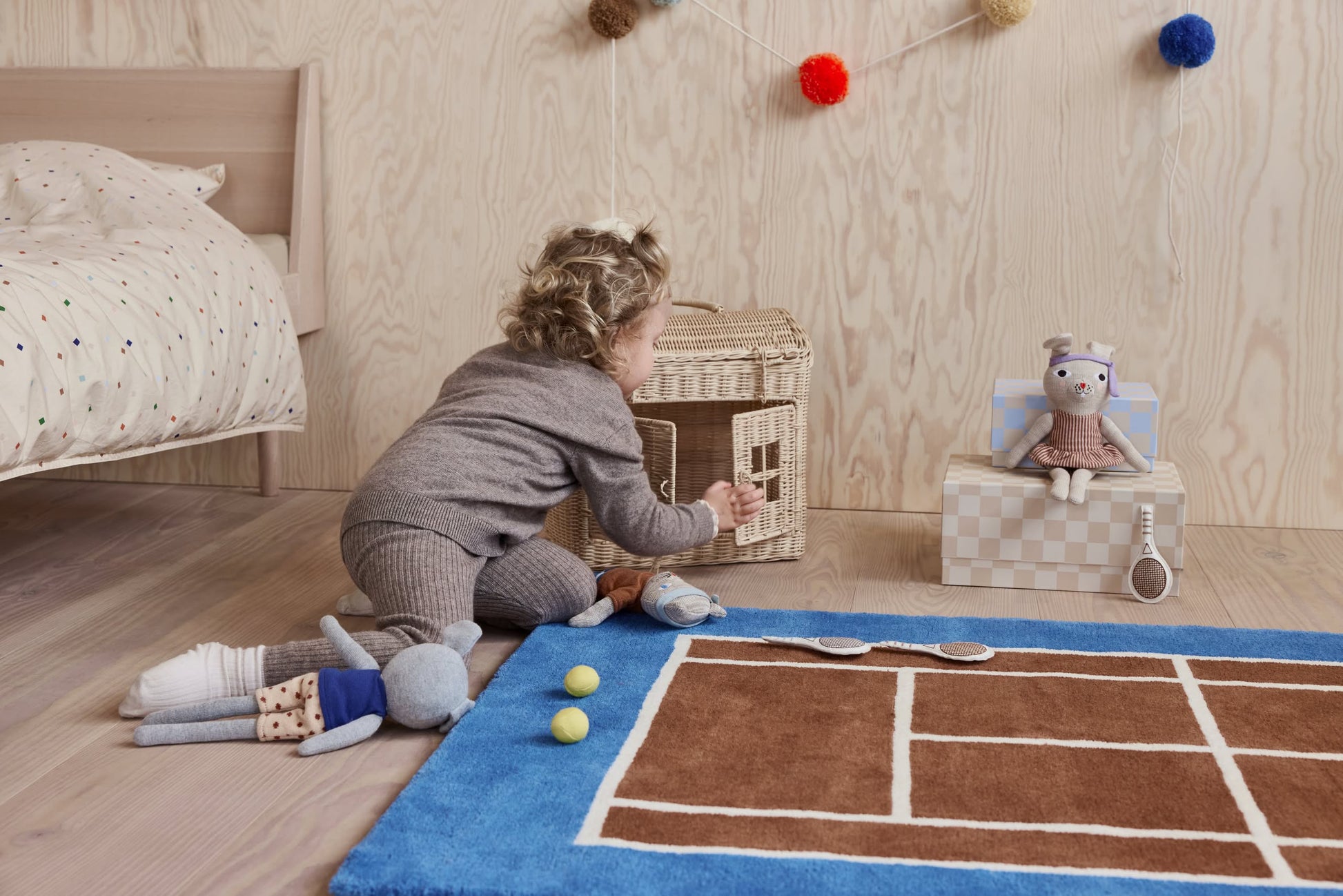 A young child in gray pajamas opens a wicker basket on the bedroom floor, where soft toys and tennis balls lie atop the OYOY Living Design Tennis Rug, adding sporty charm to the space. A plush animal rests on a checkered box in the background.