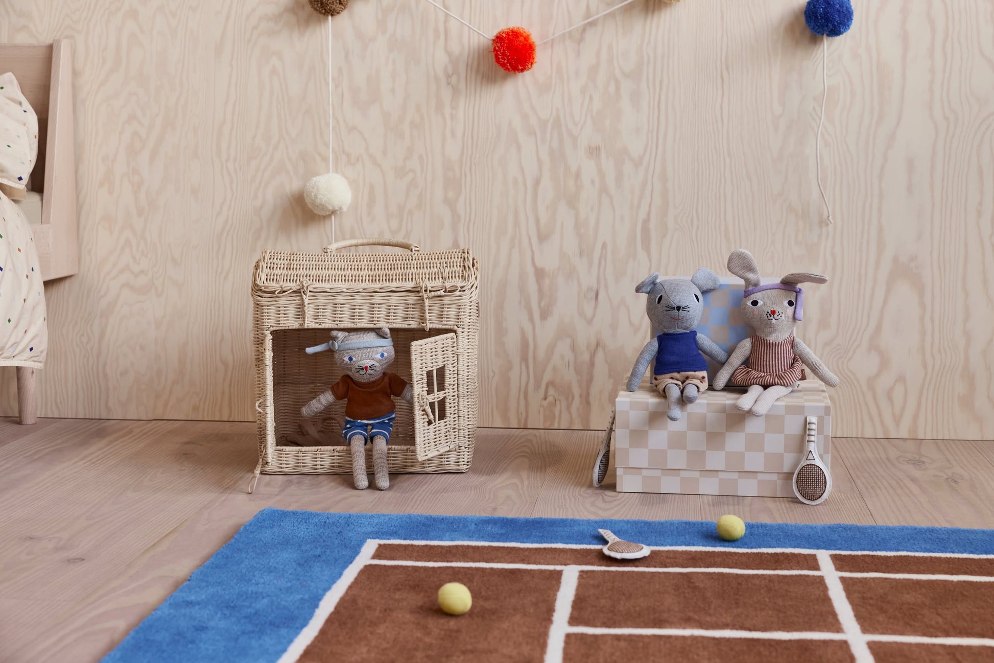 Two stuffed animals, a dog and a rabbit, sit on a checkered box by the OYOY Living Design Tennis Rug, adding playful flair. Another plush relaxes in a woven house while toy tennis rackets and balls are scattered around.