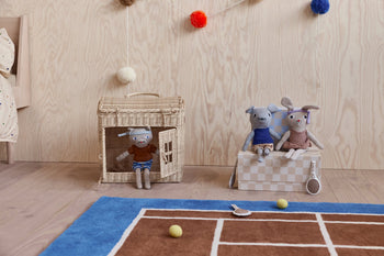 Two stuffed animals, a dog and a rabbit, sit on a checkered box by the OYOY Living Design Tennis Rug, adding playful flair. Another plush relaxes in a woven house while toy tennis rackets and balls are scattered around.