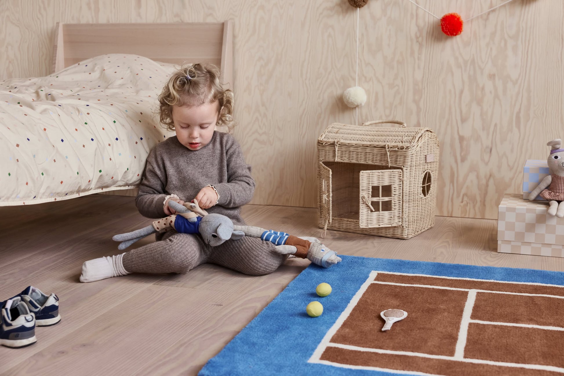 A young child plays with soft animal dolls on the OYOY Living Design Tennis Rug by OYOY Living Design, surrounded by a toy racket, two balls, a wicker house, and a pair of shoes beside the bed.