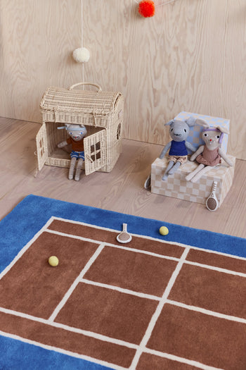 Two stuffed animal dolls sit in a small wicker house, while two others rest on a checkered box. In front is the OYOY Living Design Tennis Rug, featuring playful tennis balls and a racket on its wool and cotton-blend surface.