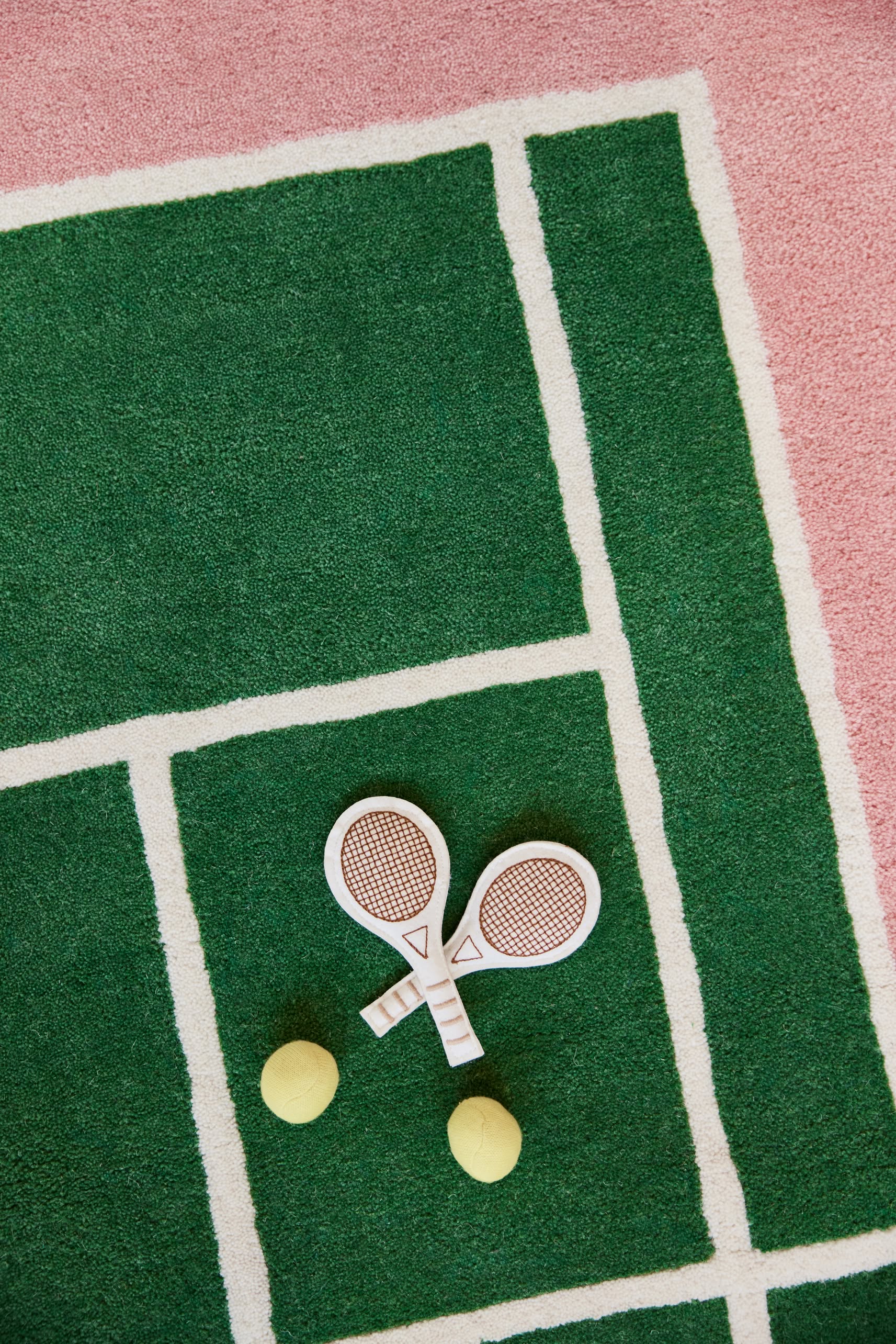 Two toy tennis rackets and yellow balls rest on the OYOY Living Design Tennis Rug, crafted by OYOY Living Design from a wool-cotton blend and featuring a sporty green and pink design inspired by real tennis courts.