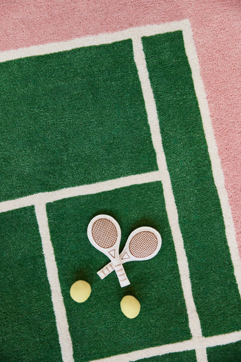 Two toy tennis rackets and yellow balls rest on the OYOY Living Design Tennis Rug, crafted by OYOY Living Design from a wool-cotton blend and featuring a sporty green and pink design inspired by real tennis courts.
