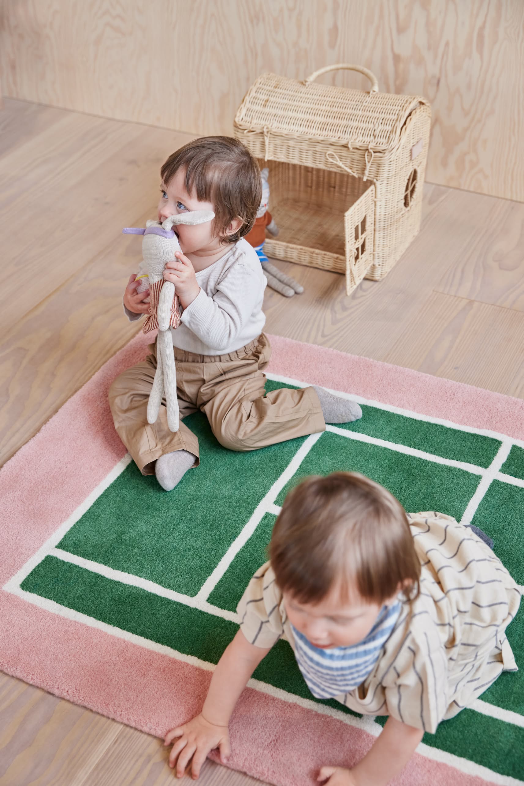Two toddlers enjoy the OYOY Living Design Tennis Rug—one sits with a stuffed toy, while the other crawls nearby. A small wicker house rests on the wooden floor, adding charm and a sporty touch to this cozy scene.