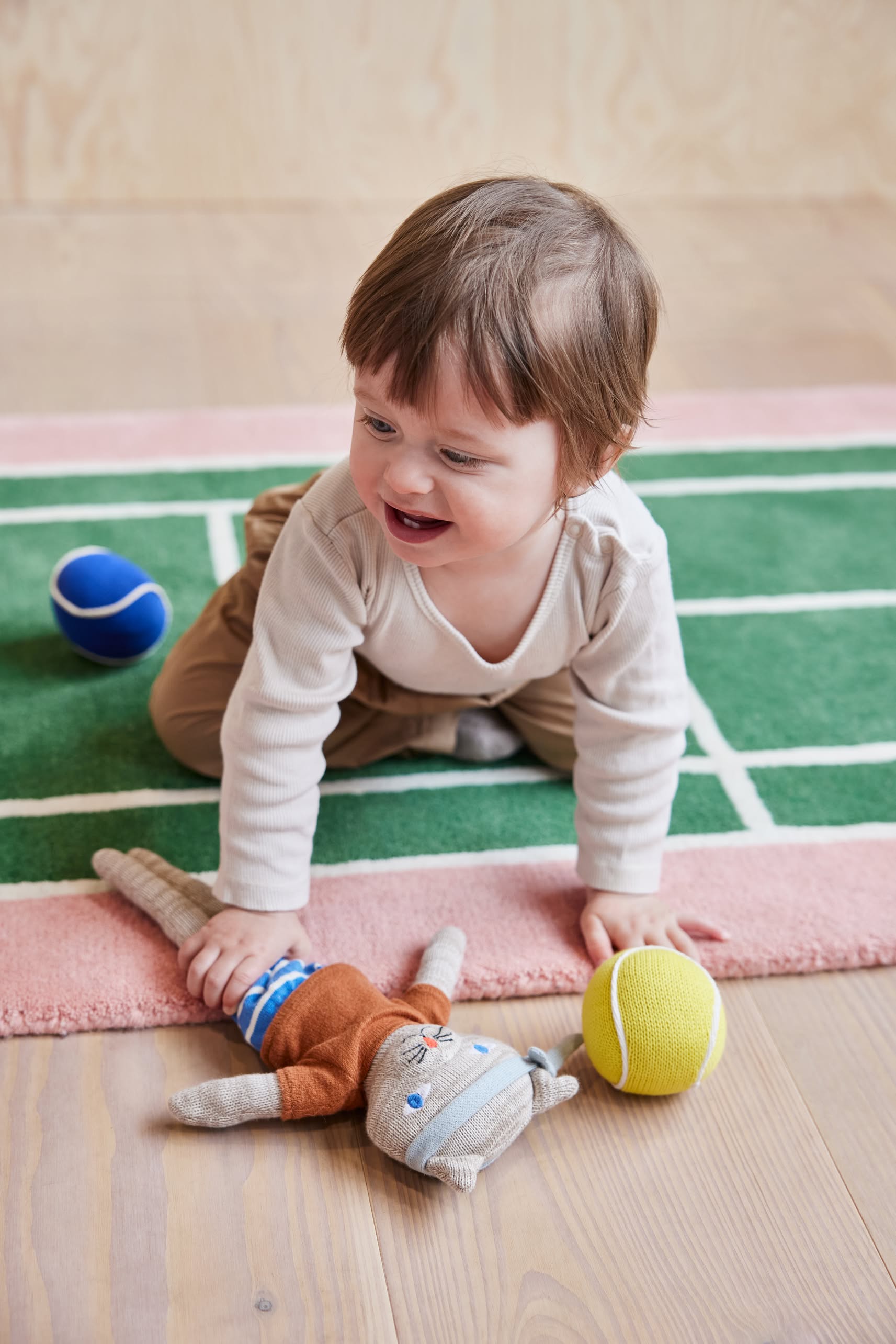 A smiling toddler in light clothes crawls on the OYOY Living Design Tennis Rug, a sporty wool-cotton blend by OYOY Living Design, with a blue ball, yellow tennis ball, and stuffed cat toy nearby.