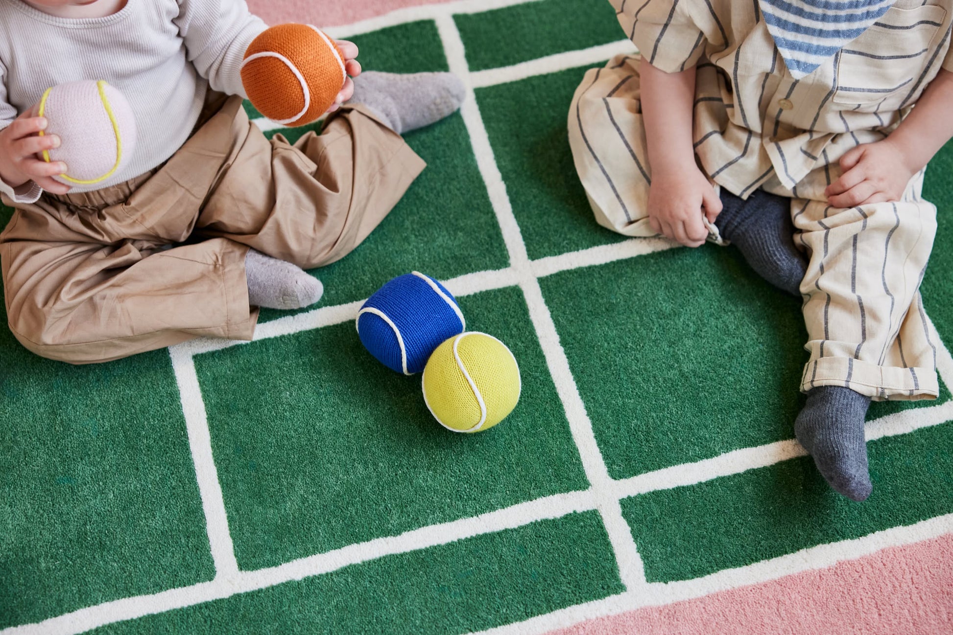 Two children sit on the OYOY Living Design Tennis Rug, a green wool and cotton blend mat with white grid lines by OYOY Living Design. They hold colorful soft toy balls, with more tennis ball-like toys scattered between them. Their faces are not visible.