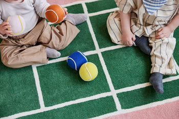 Two children sit on the OYOY Living Design Tennis Rug, a green wool and cotton blend mat with white grid lines by OYOY Living Design. They hold colorful soft toy balls, with more tennis ball-like toys scattered between them. Their faces are not visible.