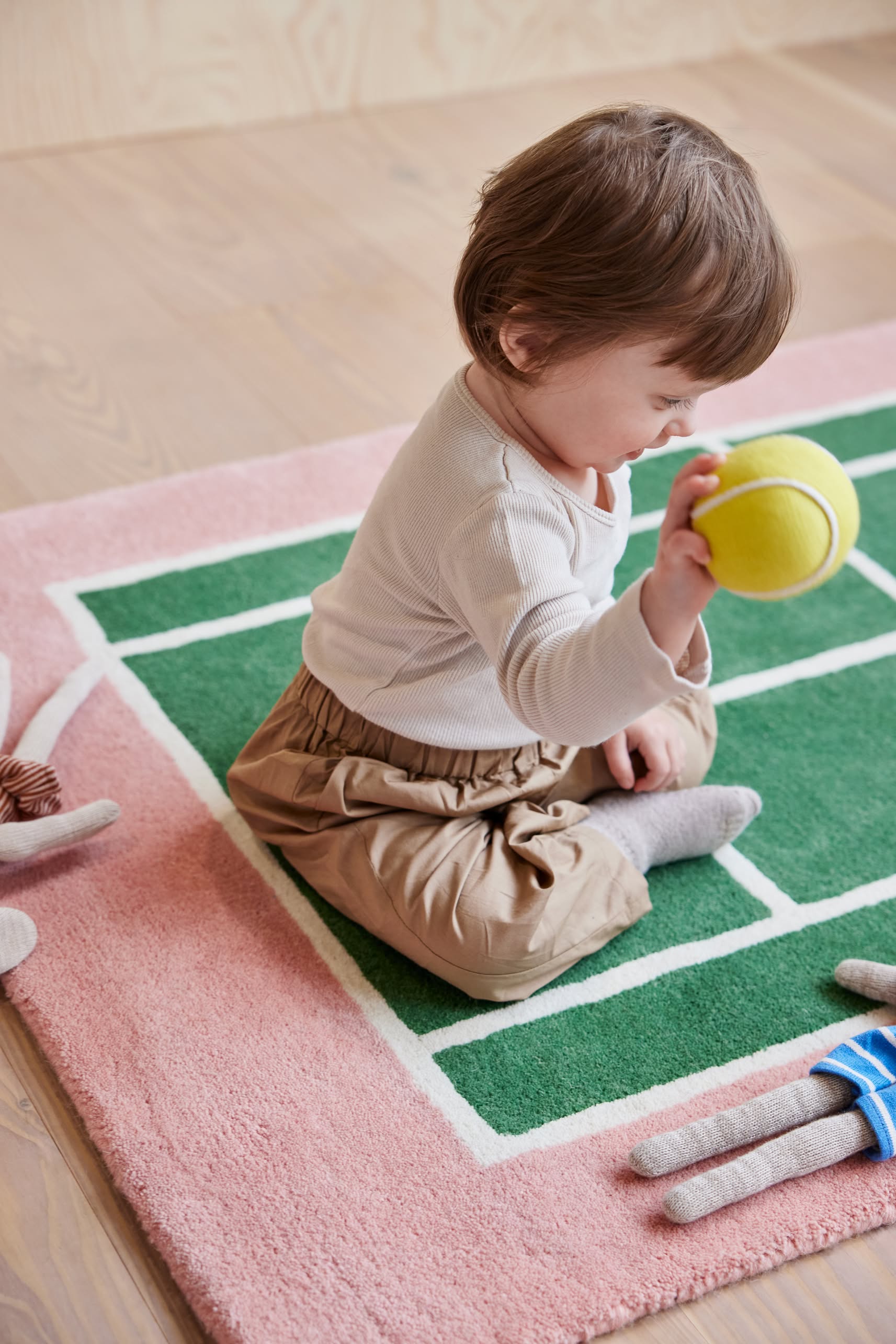 A toddler sits on the OYOY Living Design Tennis Rug by OYOY Living Design, holding a yellow tennis ball. Soft toys are nearby, and the child looks down at the ball, adding a sporty touch to this cozy scene.