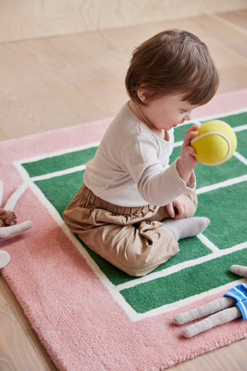 A toddler sits on the OYOY Living Design Tennis Rug by OYOY Living Design, holding a yellow tennis ball. Soft toys are nearby, and the child looks down at the ball, adding a sporty touch to this cozy scene.