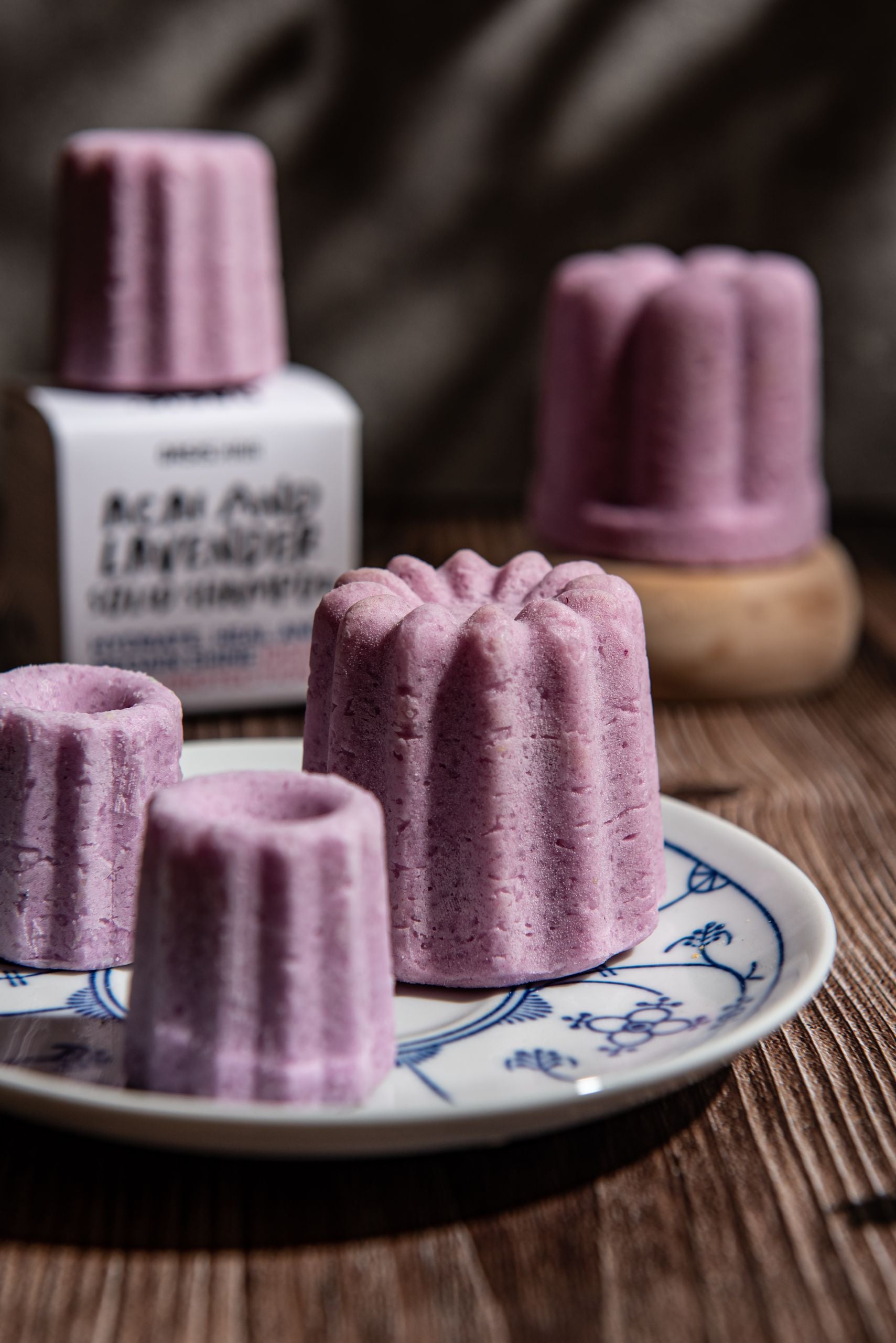 Oasis Beauty Kitchen Acai & Lavender Solid Shampoo, Midi Canele, features pink, flower-shaped bars with Organic Lavandin Oil on a patterned plate and wooden surface. A blurred shampoo bottle and natural light create a cozy, rustic atmosphere.
