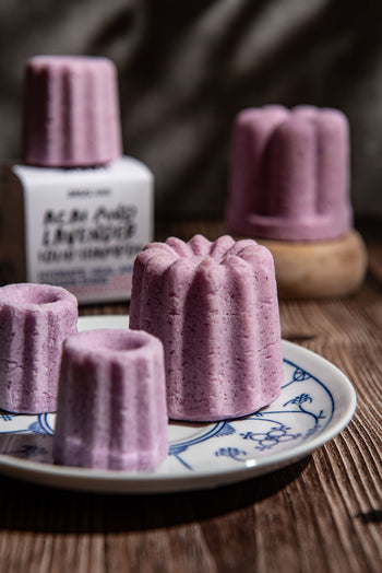 Oasis Beauty Kitchen Acai & Lavender Solid Shampoo, Midi Canele, features pink, flower-shaped bars with Organic Lavandin Oil on a patterned plate and wooden surface. A blurred shampoo bottle and natural light create a cozy, rustic atmosphere.