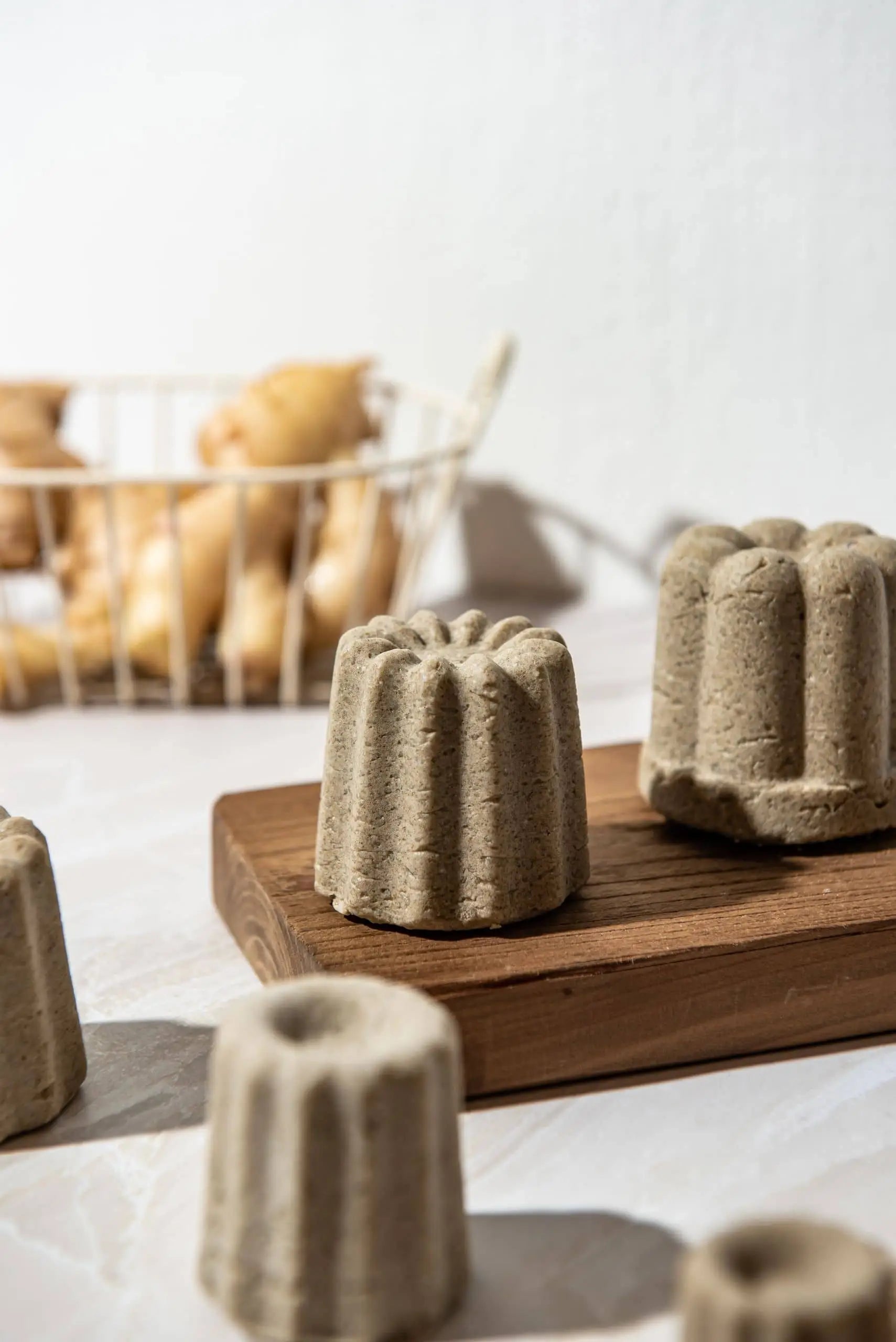Four Oasis Beauty Kitchen Ginger Solid Shampoo Midi Canele bars sit on a wooden board, with a wire basket of ginger roots—used in organic formulas for hair loss—in the background. Bright, natural lighting highlights their textured surface.
