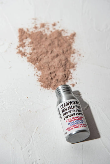 A small open bottle labeled Oasis Beauty Kitchen Head to Toe Magic Powder Wash, 25g lies on a white surface with a pile of pinkish powder, made from plant-based ingredients and organic essential oils, spilled behind it. Suitable for face, hair, and body care.